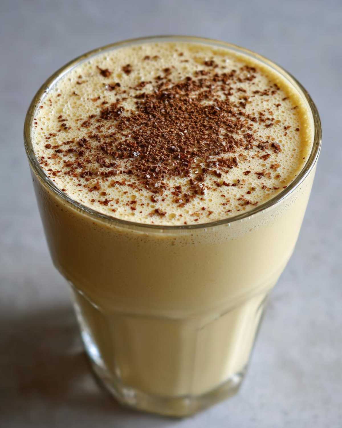 Close-up of a tall glass filled with creamy, yellow eggnog, topped with foamy froth and grated nutmeg.