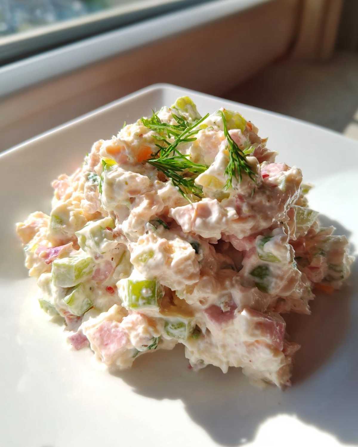 A close-up serving of creamy ham salad recipe mixed with celery and topped with fresh dill, sitting on a white plate.
