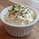 A small white ramekin filled with thick, creamy horseradish sauce, garnished with fresh chopped chives.