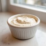 A small white ramekin filled with creamy, slightly speckled horseradish sauce, set on a white marble surface.