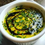 Close-up of a creamy olive oil bread dip, bright yellow with chopped green herbs and a balsamic glaze swirl.