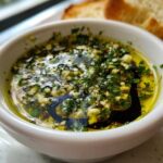 Close-up of a white bowl containing olive oil bread dip with visible minced garlic, herbs, and balsamic vinegar, served with bread slices.