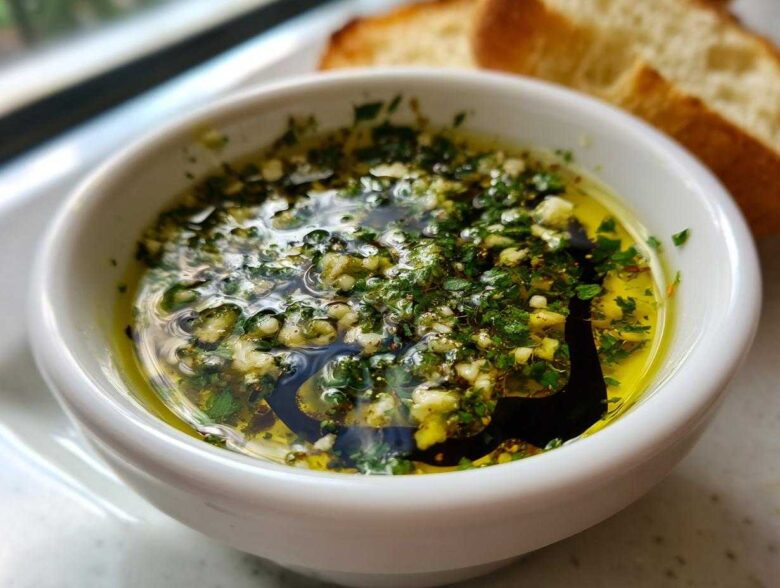 Close-up of a white bowl containing olive oil bread dip with visible minced garlic, herbs, and balsamic vinegar, served with bread slices.