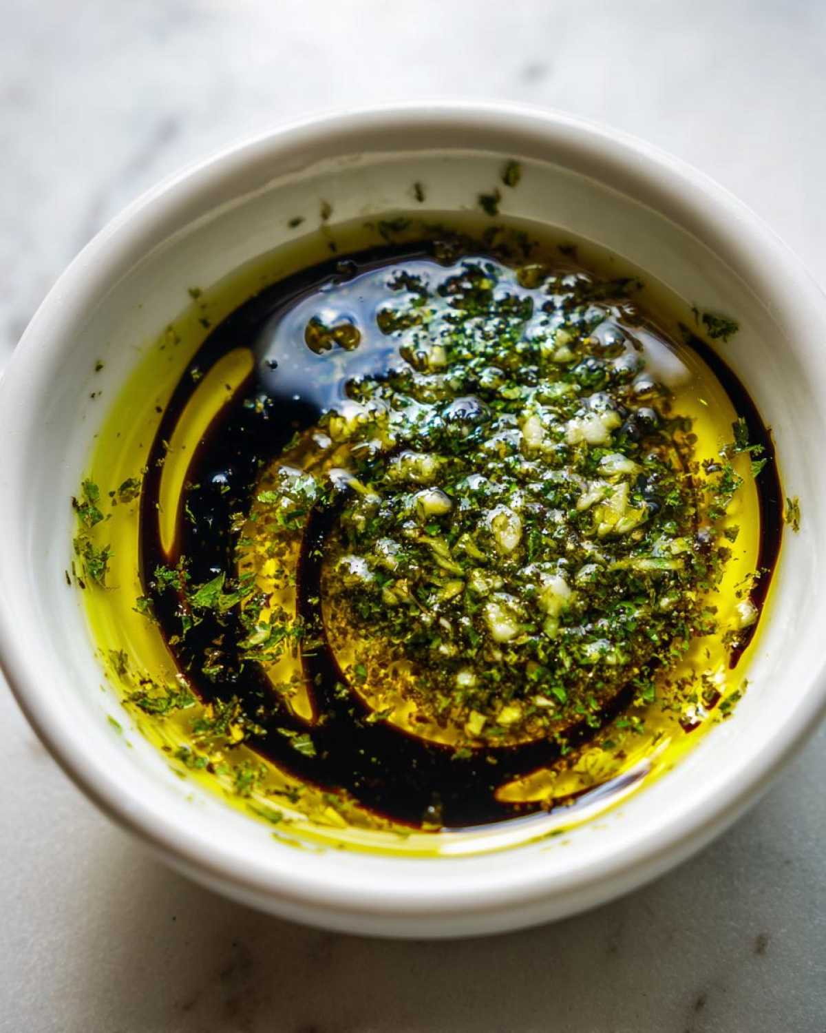 Close-up of a creamy olive oil bread dip in a white bowl, featuring bright green herbs, garlic, and a swirl of dark balsamic vinegar.