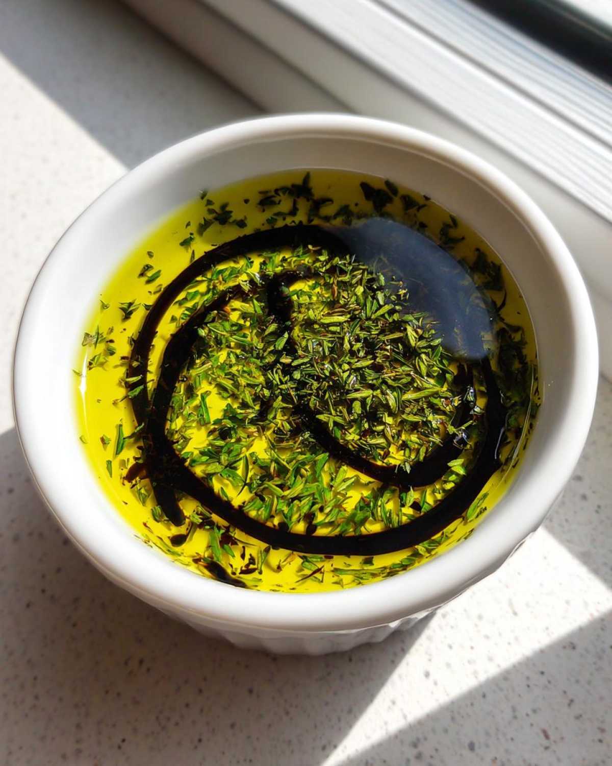 Close-up of a white bowl containing olive oil bread dip, topped with fresh herbs and a swirl of balsamic vinegar.