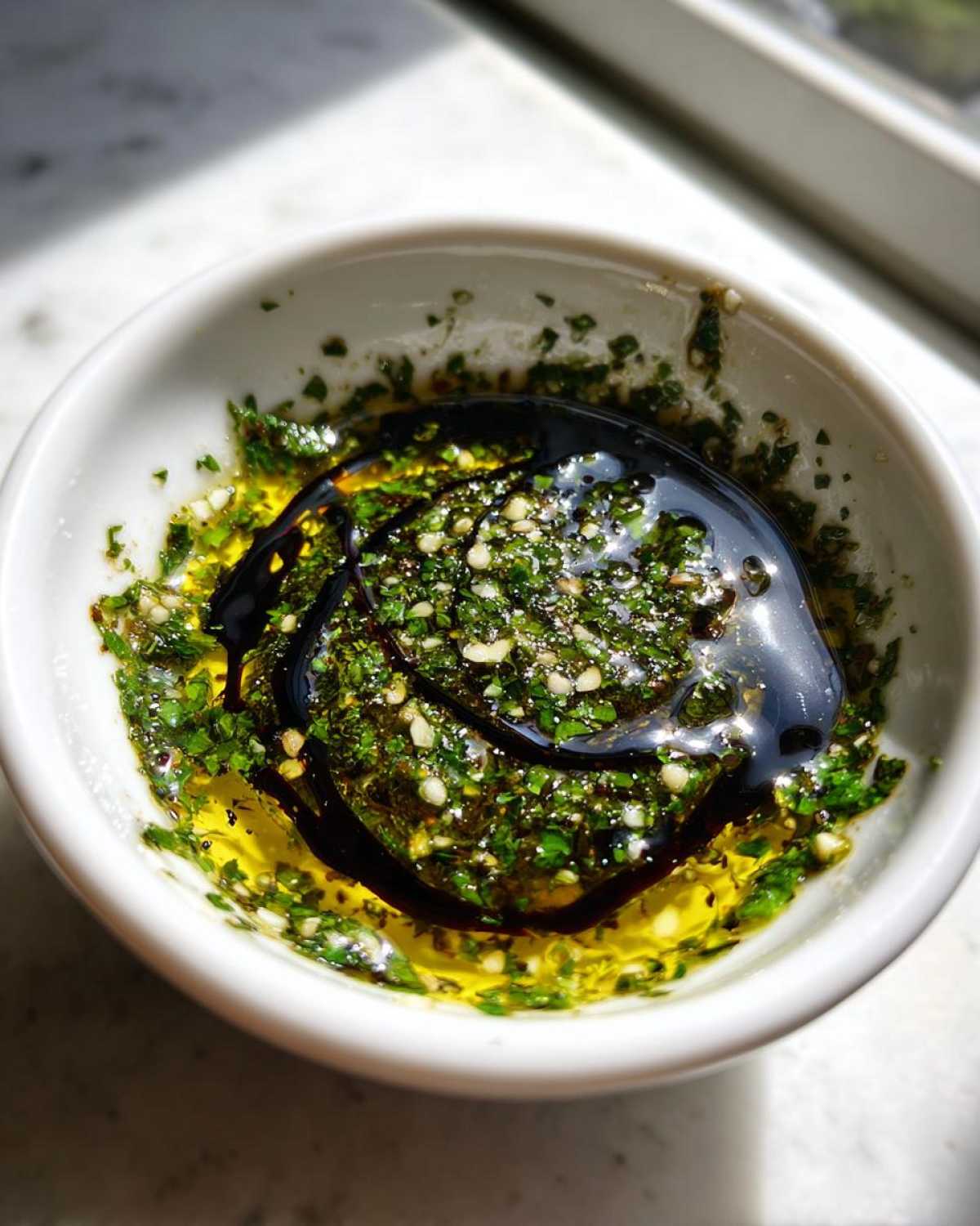 Close-up of olive oil bread dip ingredients: olive oil, herbs, garlic, and balsamic glaze in a white bowl.