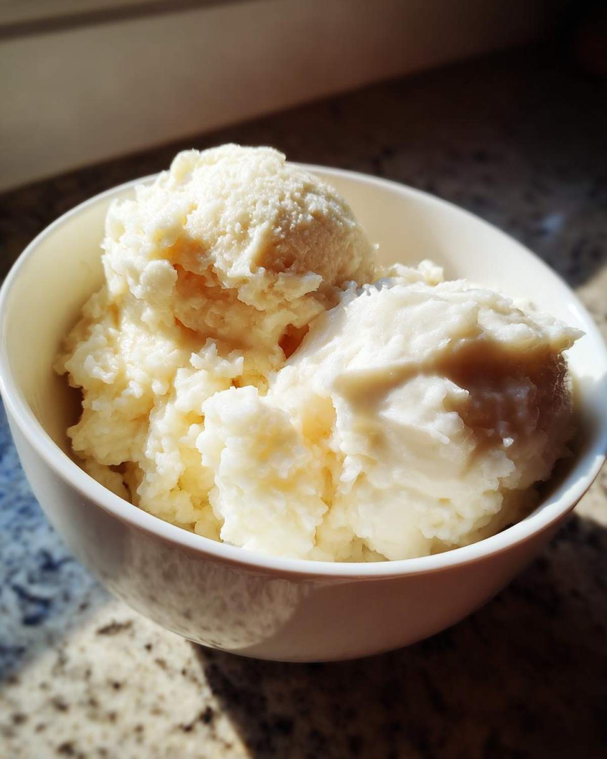 Two generous scoops of creamy, white rice pudding served in a white bowl, catching bright sunlight.
