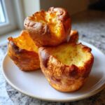 Three golden brown, crispy yorkshire pudding puffs stacked slightly on a light-colored plate.