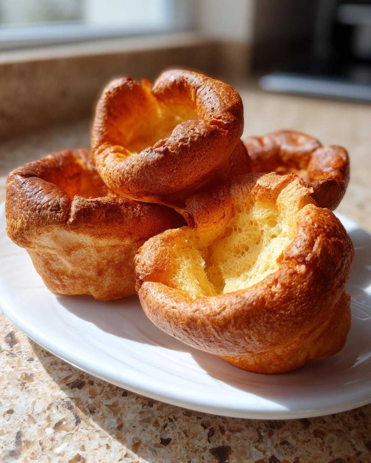 A close-up of four golden brown, crispy yorkshire pudding puffs stacked slightly on a white plate.