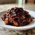 A mound of tender, shredded crock pot pulled pork generously coated in dark, glossy barbecue sauce on a white plate.