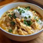 A close-up of a bowl of rich crockpot white chicken chili, topped with sour cream and fresh cilantro.