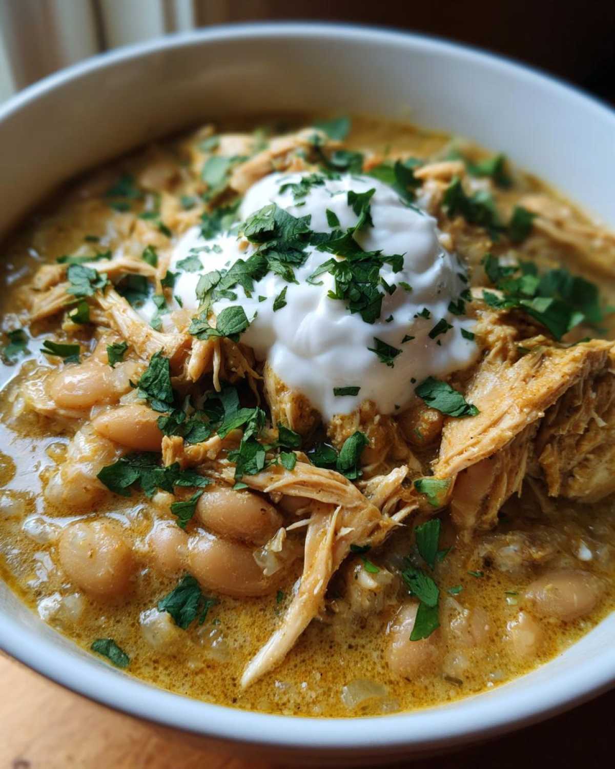 Close-up of a bowl of creamy crockpot white chicken chili topped with shredded chicken, sour cream, and fresh cilantro.