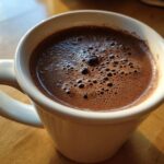 Close-up of a white mug filled with rich, foamy hot cocoa mix, sitting on a wooden table.