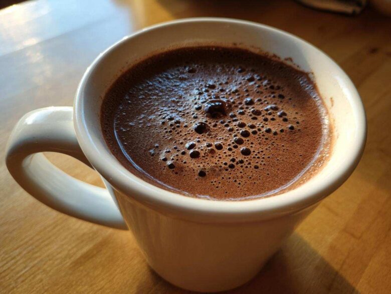 Close-up of a white mug filled with rich, foamy hot cocoa mix, sitting on a wooden table.