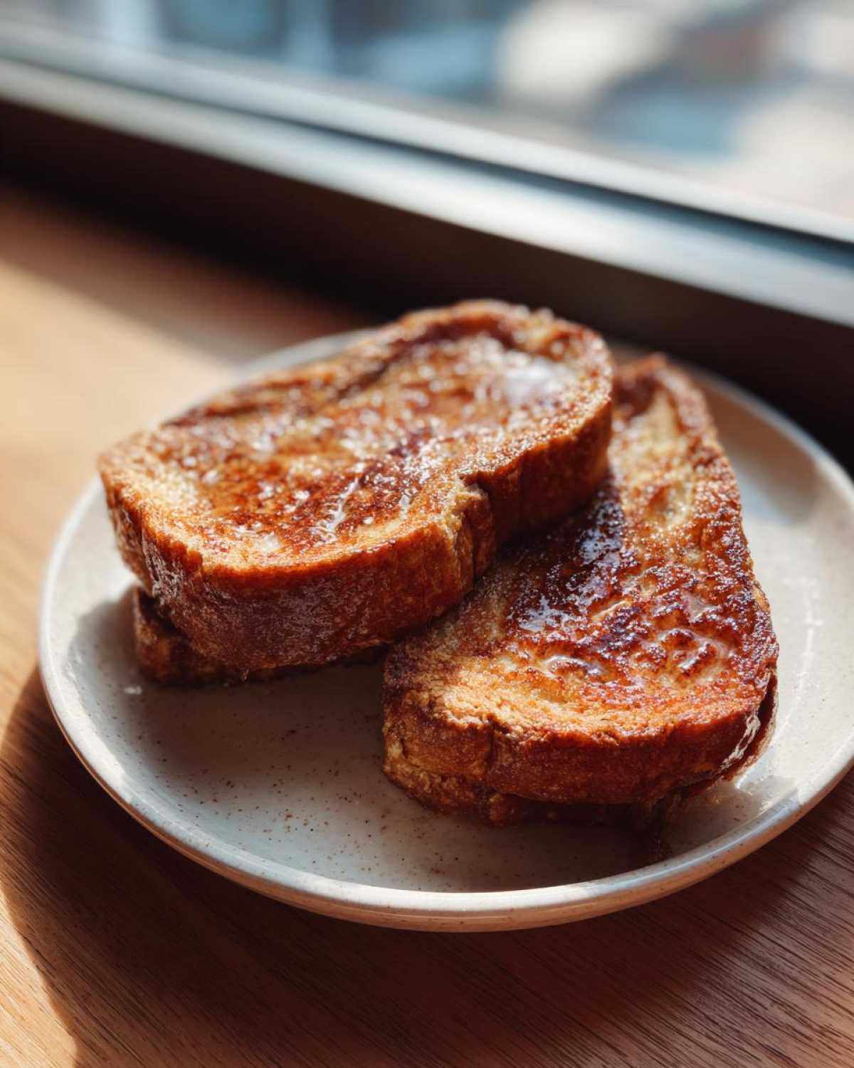 Two golden brown slices of easy french toast drizzled with syrup, resting on a speckled white plate near a window.