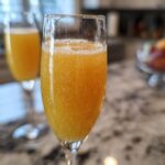 Close-up of a fizzy, orange bellini recipe cocktail served in a champagne flute glass on a marble countertop.