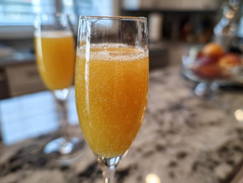 Close-up of a fizzy, orange bellini recipe cocktail served in a champagne flute glass on a marble countertop.
