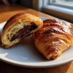 Two golden brown chocolate croissants on a white plate, one cut open to show melted chocolate filling.