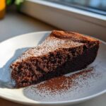 A rich slice of flourless chocolate torte dusted with powdered sugar and cocoa powder on a white plate.