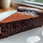 A decadent slice of flourless chocolate torte dusted with cocoa powder and powdered sugar, resting on a white plate.