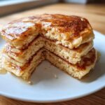 A stack of three fluffy almond flour pancakes drizzled generously with maple syrup, showing a bite taken out.