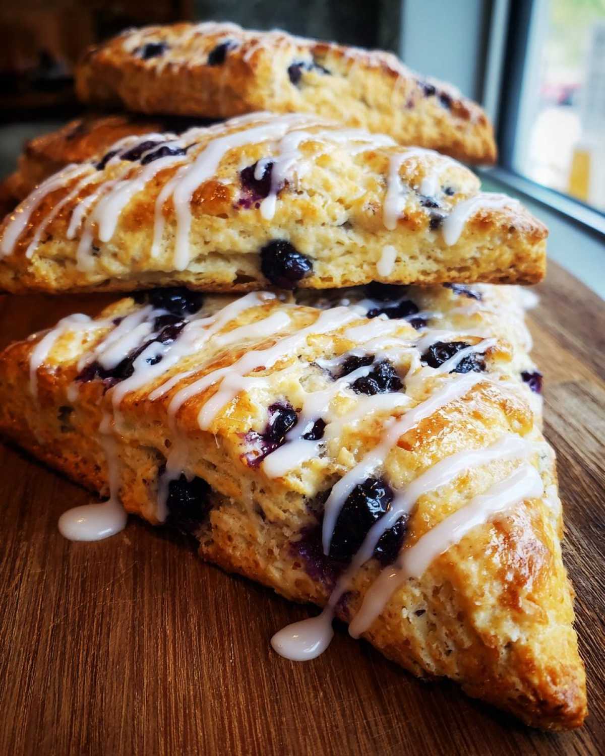 A stack of three freshly baked blueberry scones topped with a thick white vanilla glaze, sitting on a wooden board.
