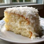 A close-up of a moist slice of fluffy coconut cake layered with white frosting and topped with shredded coconut.
