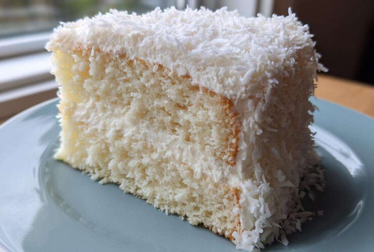 A moist slice of fluffy coconut cake covered in white frosting and shredded coconut flakes.