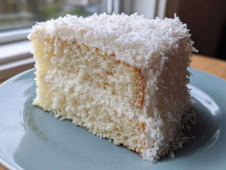 A moist slice of fluffy coconut cake covered in white frosting and shredded coconut flakes.