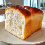 A close-up of a thick slice of soft, fluffy condensed milk bread with a shiny, golden-brown crust.