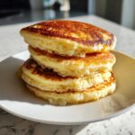 A close-up stack of four golden brown, incredibly fluffy pancakes on a white plate.
