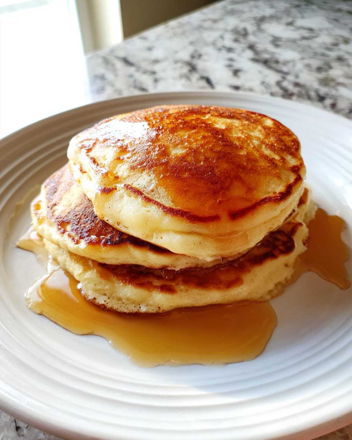 A stack of three fluffy sourdough pancakes drizzled generously with golden maple syrup on a white plate.