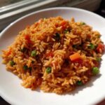 A close-up of fluffy, orange-red spanish rice mixed with bright green peas and diced carrots served in a white bowl.