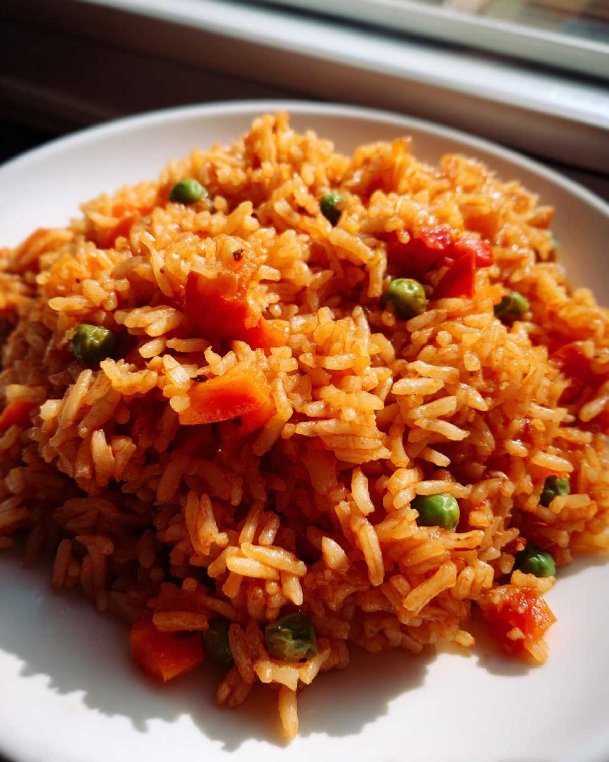 Close-up of fluffy, orange-colored spanish rice mixed with bright green peas and diced carrots on a white plate.