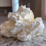 A close-up of a fluffy, pale yellow scoop of homemade vanilla buttercream frosting resting on a granite countertop.