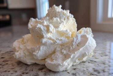 A close-up of a fluffy, pale yellow scoop of homemade vanilla buttercream frosting resting on a granite countertop.