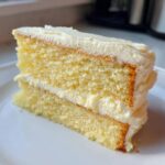 A close-up of a fluffy, two-layer slice of vanilla cake recipe covered in white buttercream frosting.