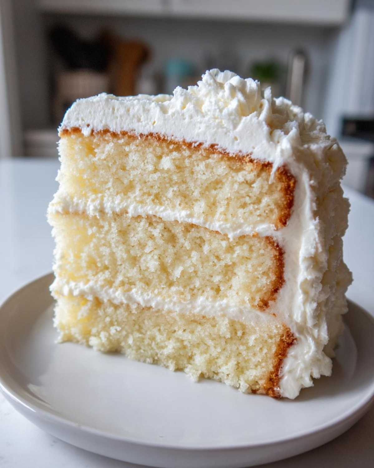Close-up of a moist, three-layer slice of vanilla cake recipe covered in thick white frosting.