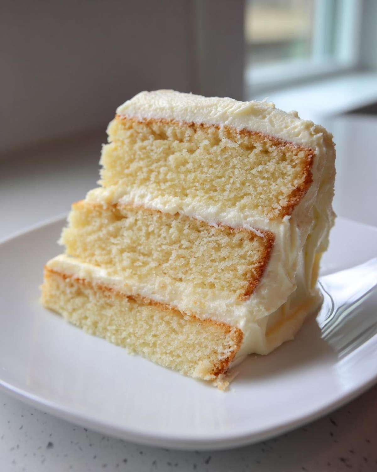 Close-up of a moist, three-layer slice of vanilla cake recipe covered in creamy white frosting on a white plate.