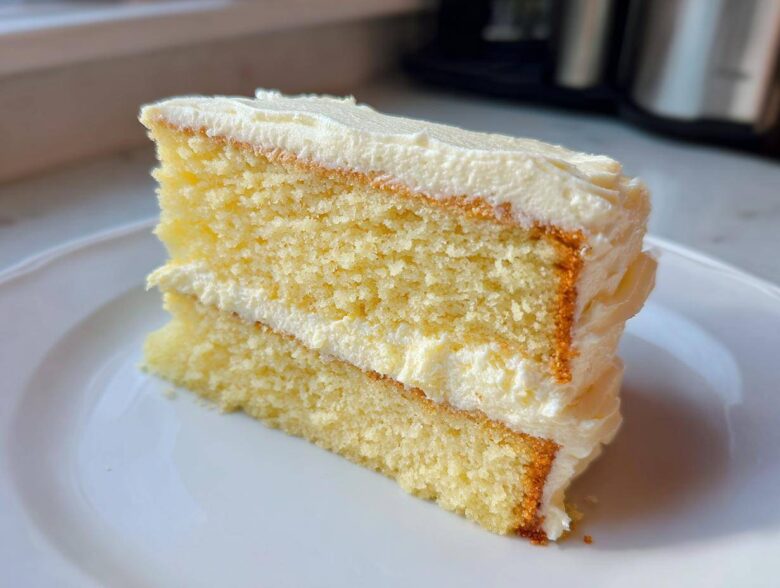 A close-up of a fluffy, two-layer slice of vanilla cake recipe covered in white buttercream frosting.