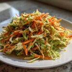 A mound of bright, shredded cabbage salad mixed with julienned carrots and herbs, served on a white plate.