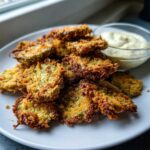 A pile of crispy, golden brown Fried Pickles served on a white plate next to a small bowl of creamy dipping sauce.