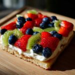 Close-up of a slice of fruit pizza sugar cookie topped with whipped cream, strawberries, blueberries, and kiwi slices.