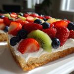 A close-up of a slice of fruit pizza sugar cookie topped with whipped cream, strawberries, blueberries, and kiwi.