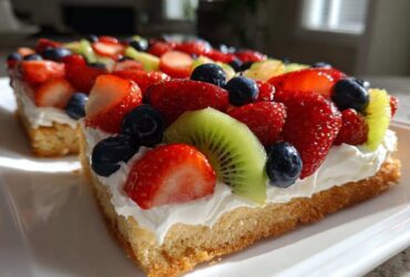 A close-up of a slice of fruit pizza sugar cookie topped with whipped cream, strawberries, blueberries, and kiwi.