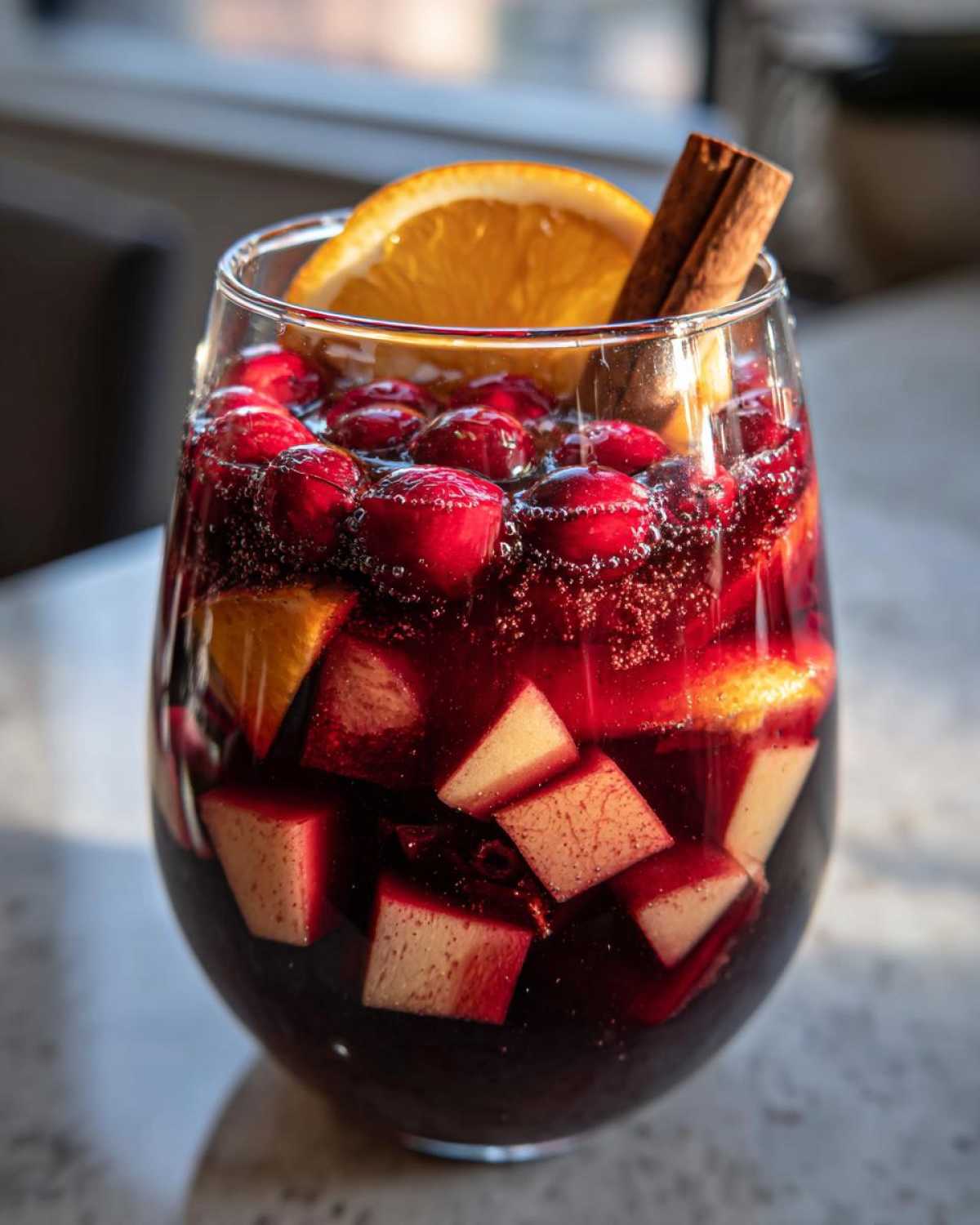 Close-up of a glass filled with vibrant Christmas sangria, featuring cranberries, apple chunks, an orange slice, and a cinnamon stick.