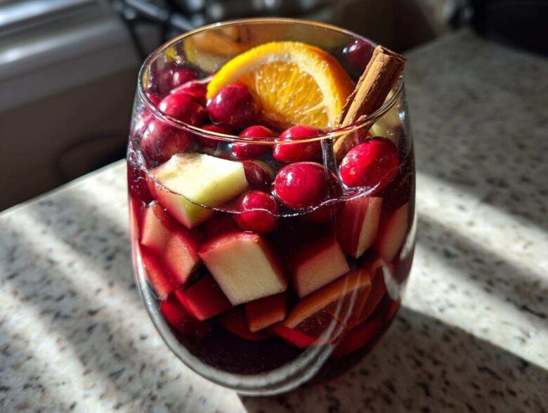 A close-up of a glass filled with rich red Christmas sangria, loaded with cranberries, apple chunks, and garnished with an orange slice and cinnamon stick.