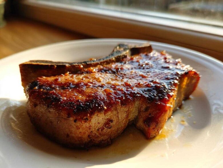 A thick, bone-in pork chop, glistening with a dark glaze, resting on a white plate.