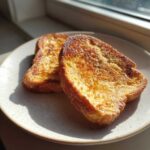 Two perfectly golden brown slices of easy french toast resting on a speckled white plate near a window.