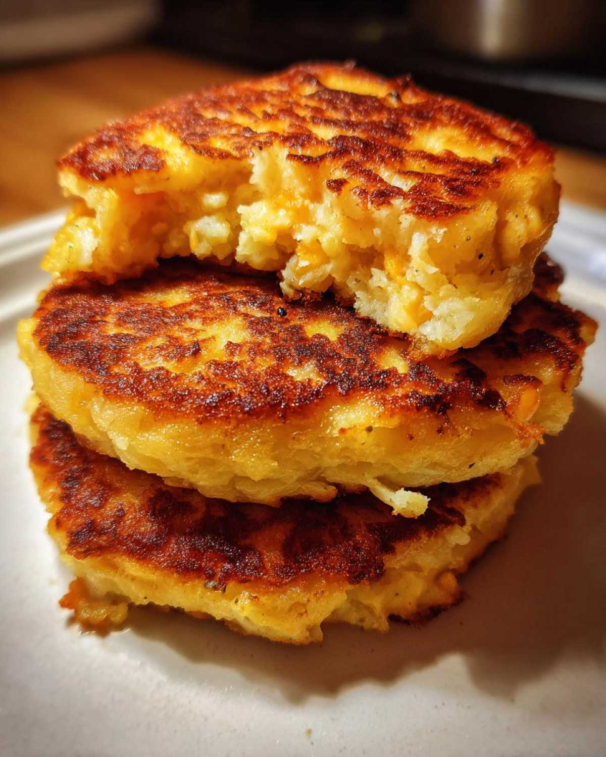 A stack of three golden-brown, pan-fried mashed potato cakes, with the top one broken open to show the texture.